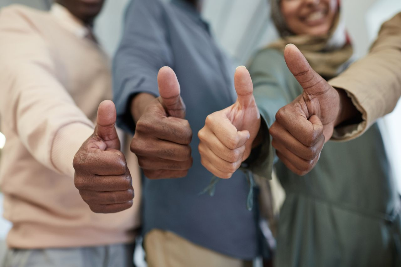 ours-journey Close-up of a diverse team giving thumbs up indoors, symbolizing success and unity.